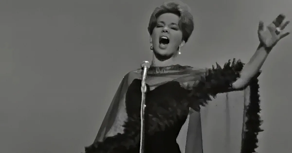 A woman in a dramatic pose, wearing a feathered cape, sings passionately into a microphone.