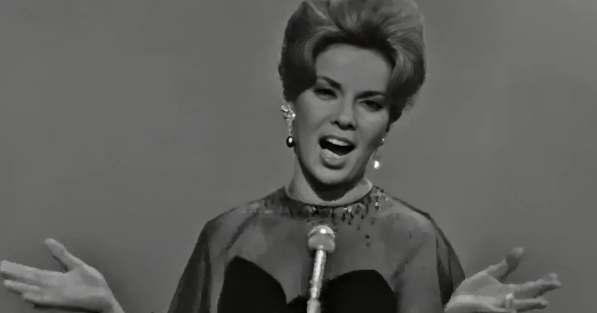 A woman in a vintage outfit sings passionately, gesturing with her hands, wearing large earrings.