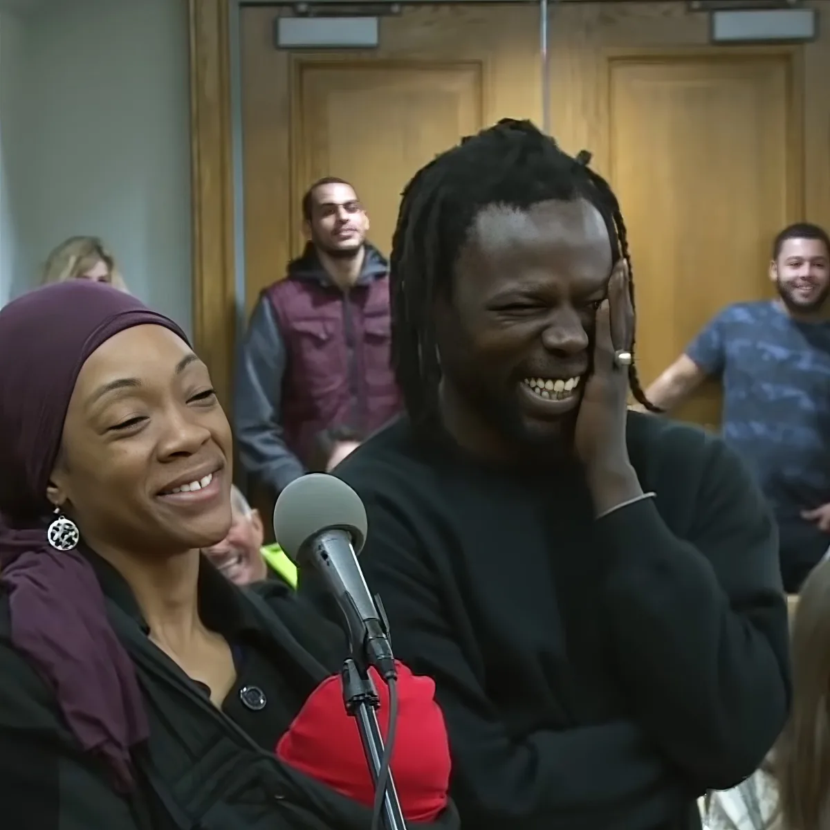 A man with dreadlocks laughs while a woman in a hijab smiles nearby, both in front of a microphone.