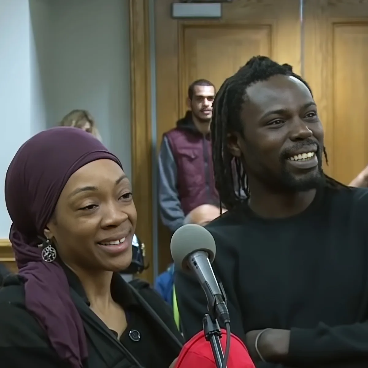 A smiling woman in a purple headscarf and a man with dreadlocks are seated in front of a microphone, likely in a studio...