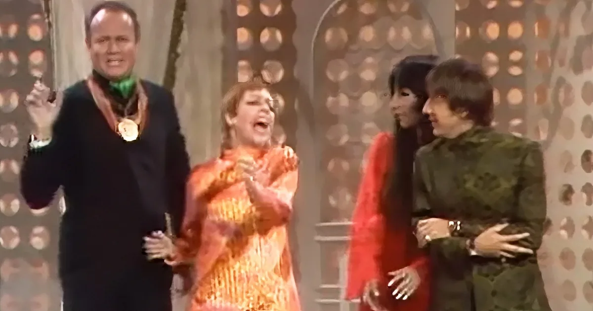 A woman in a vibrant orange dress is singing passionately on stage, surrounded by other performers.