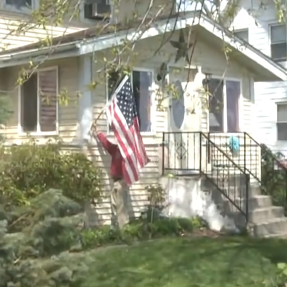 A person in red holds an American flag on a porch with a white house and greenery.