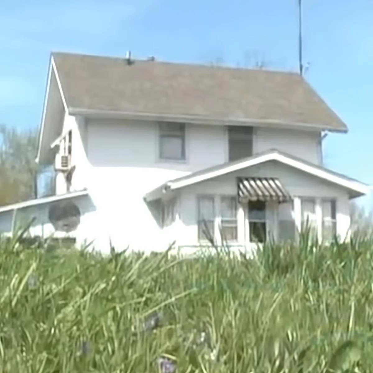A white house with a brown roof stands amidst tall grass under a clear blue sky.