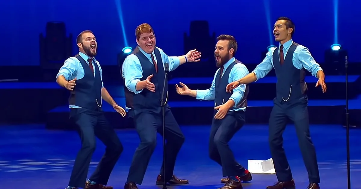 Four men in blue vests and shirts perform on stage with microphones and blue lighting.