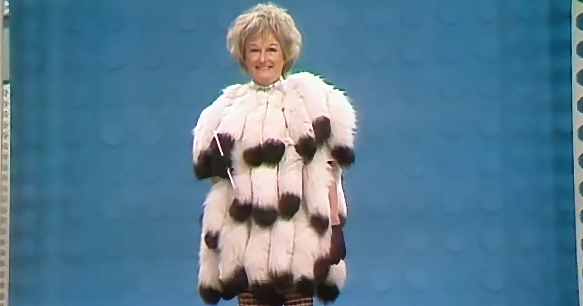 A person in a white fur coat with black accents stands against a blue background.