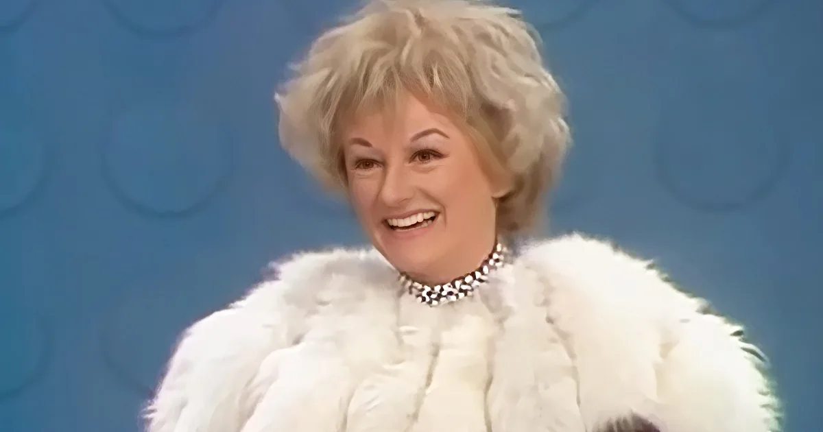 A smiling woman with blonde hair, wearing a white fur coat and pearl necklace, against a blue background.