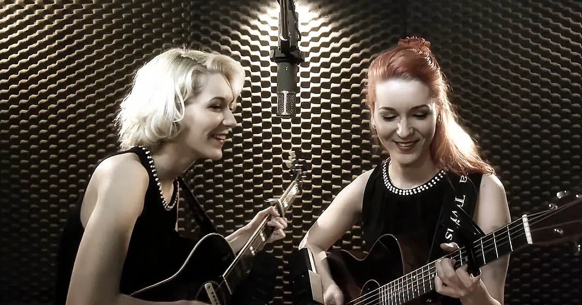 Two women in black dresses play guitars in a recording studio with acoustic foam.