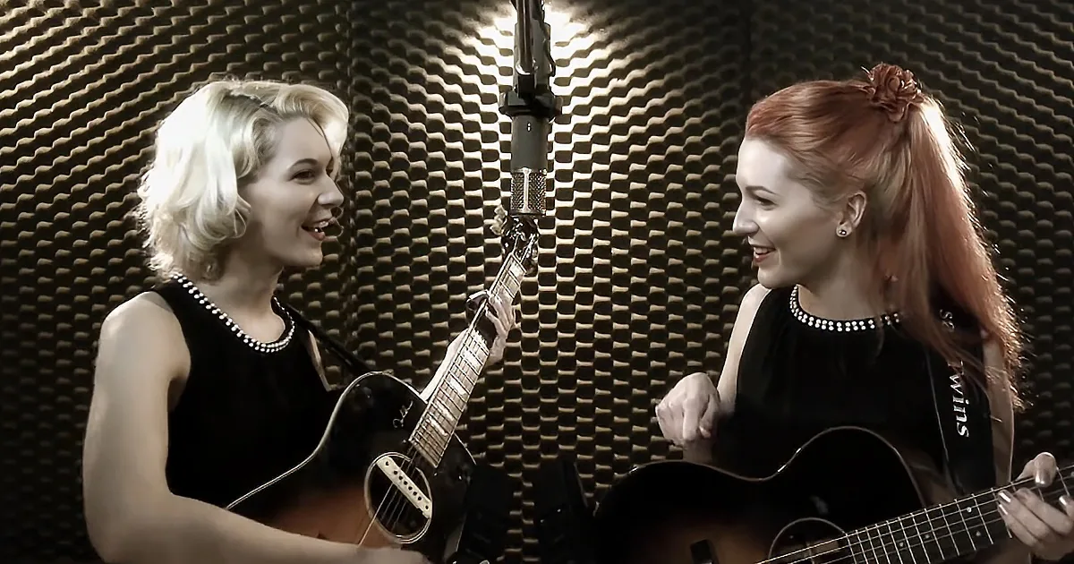Two women in a recording studio, one with blonde hair and the other with red, both playing guitars and smiling.