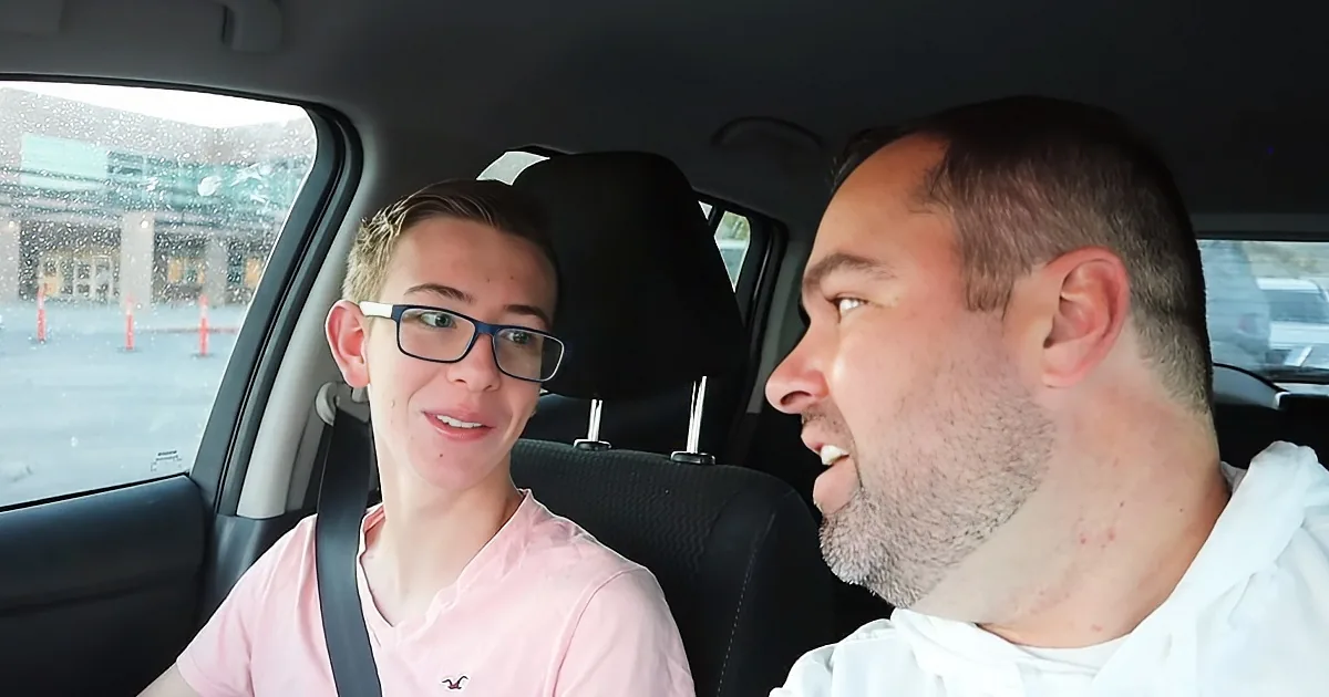A man and a boy are sitting in a car, both smiling, with the boy wearing glasses and a pink shirt.