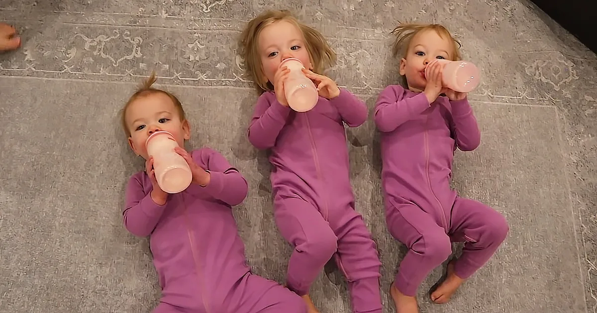 Three children in purple outfits are lying on a carpet, each drinking from a pink bottle.