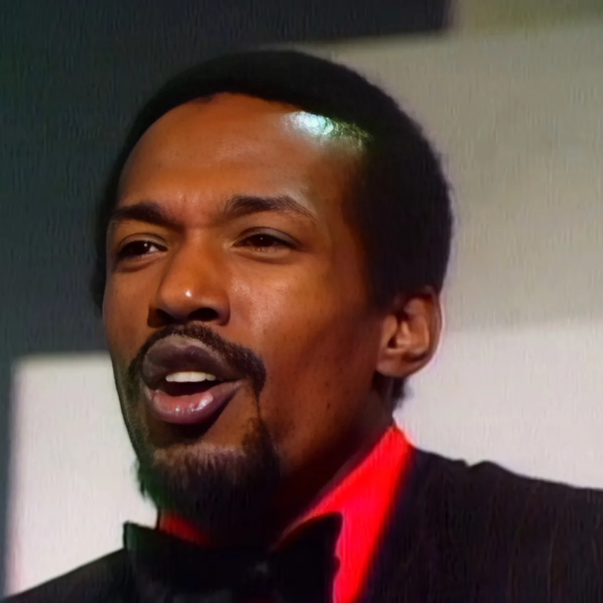 A man with short black hair and a beard, wearing a black jacket with a red collar, is speaking.