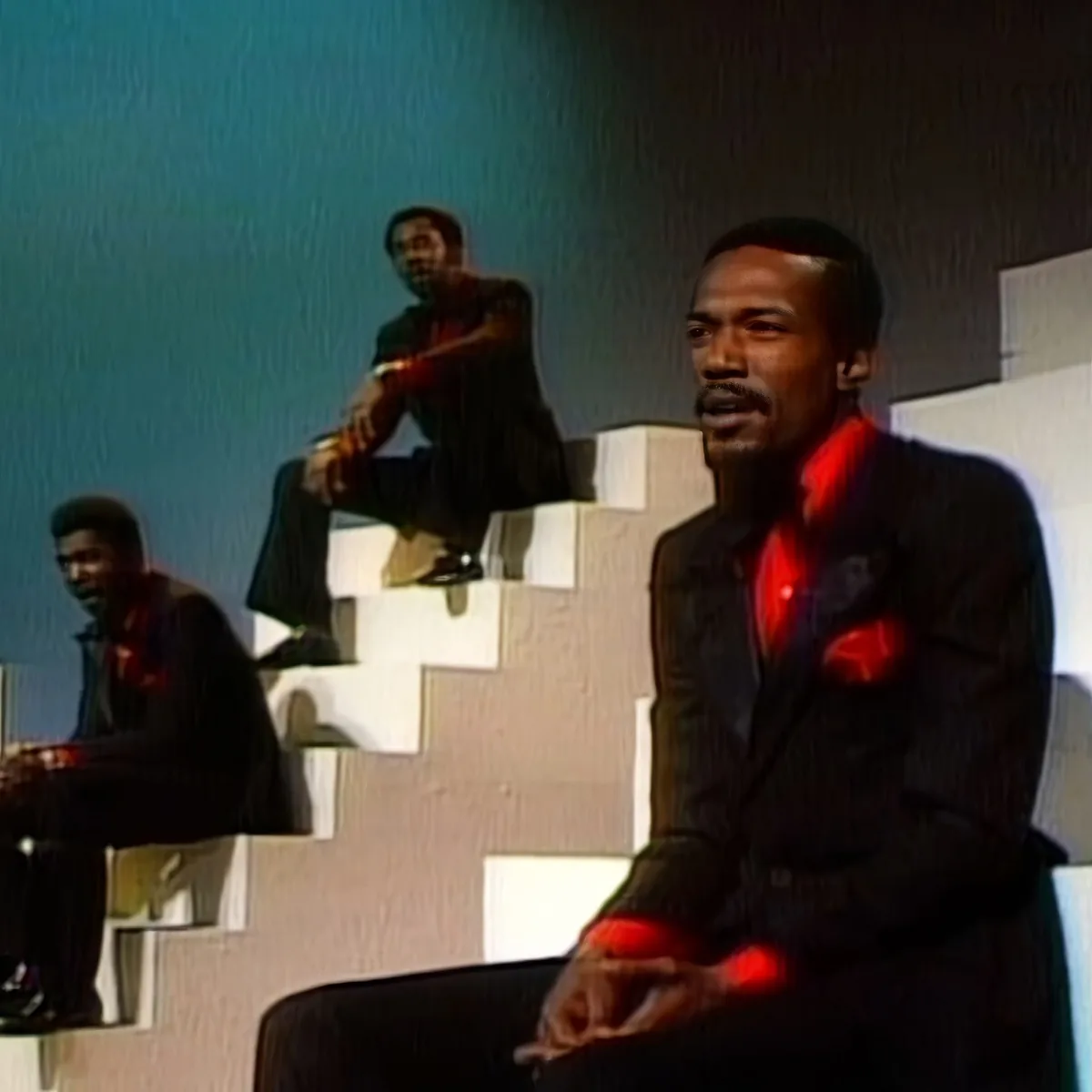 A man in a suit sits on a chair, holding a red object, with two other men in similar attire behind him.