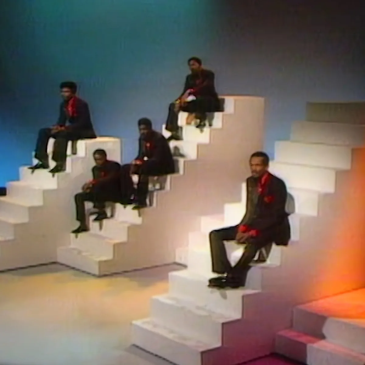 Six men in black suits with red accents sit on white steps against a gradient blue and orange background.
