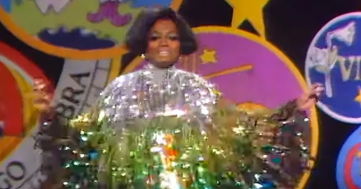 A person in a shiny, reflective outfit stands on a stage with colorful, cartoonish background.