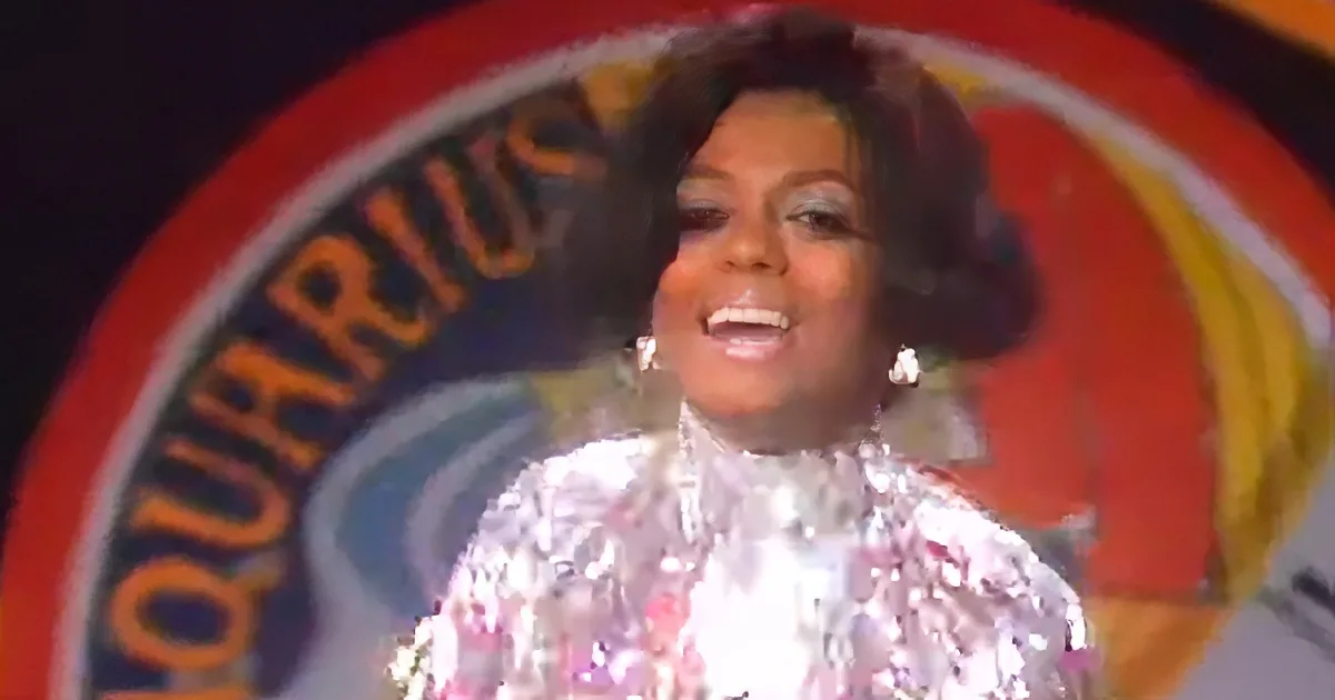 A person in a shiny silver outfit smiles in front of a colorful "Aquarius" sign.
