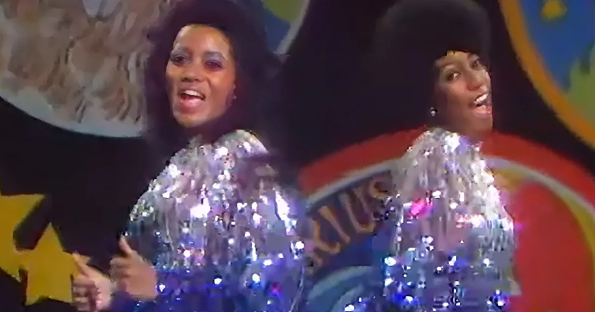 Two performers in shiny, sequined outfits sing on stage with colorful background.