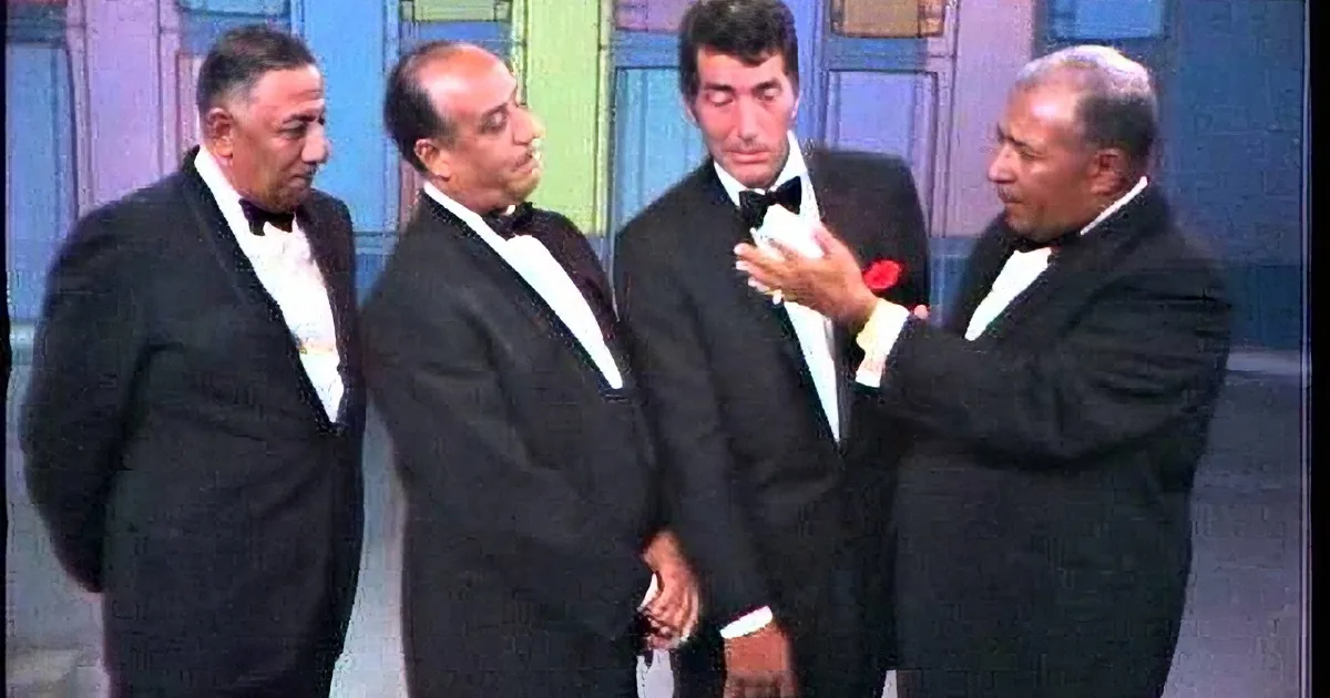 Four men in tuxedos smile and pose together on a stage with colorful background.