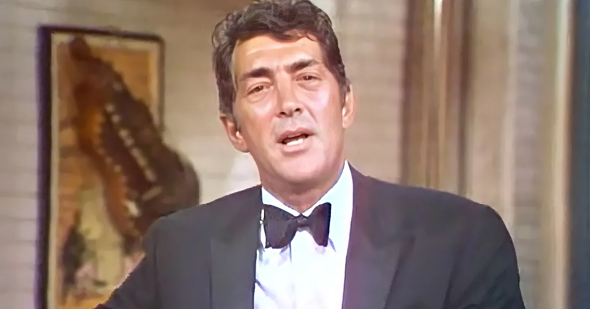 A man in a tuxedo with a bow tie is speaking, with a butterfly painting in the background.