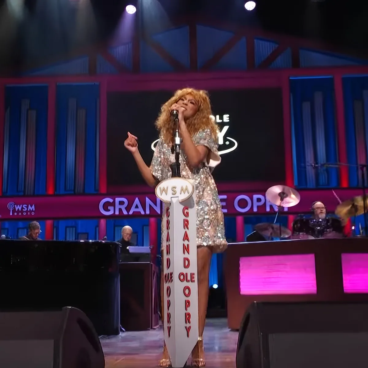 A performer in a glittery dress sings at the Grand Ole Opry stage with a band behind.