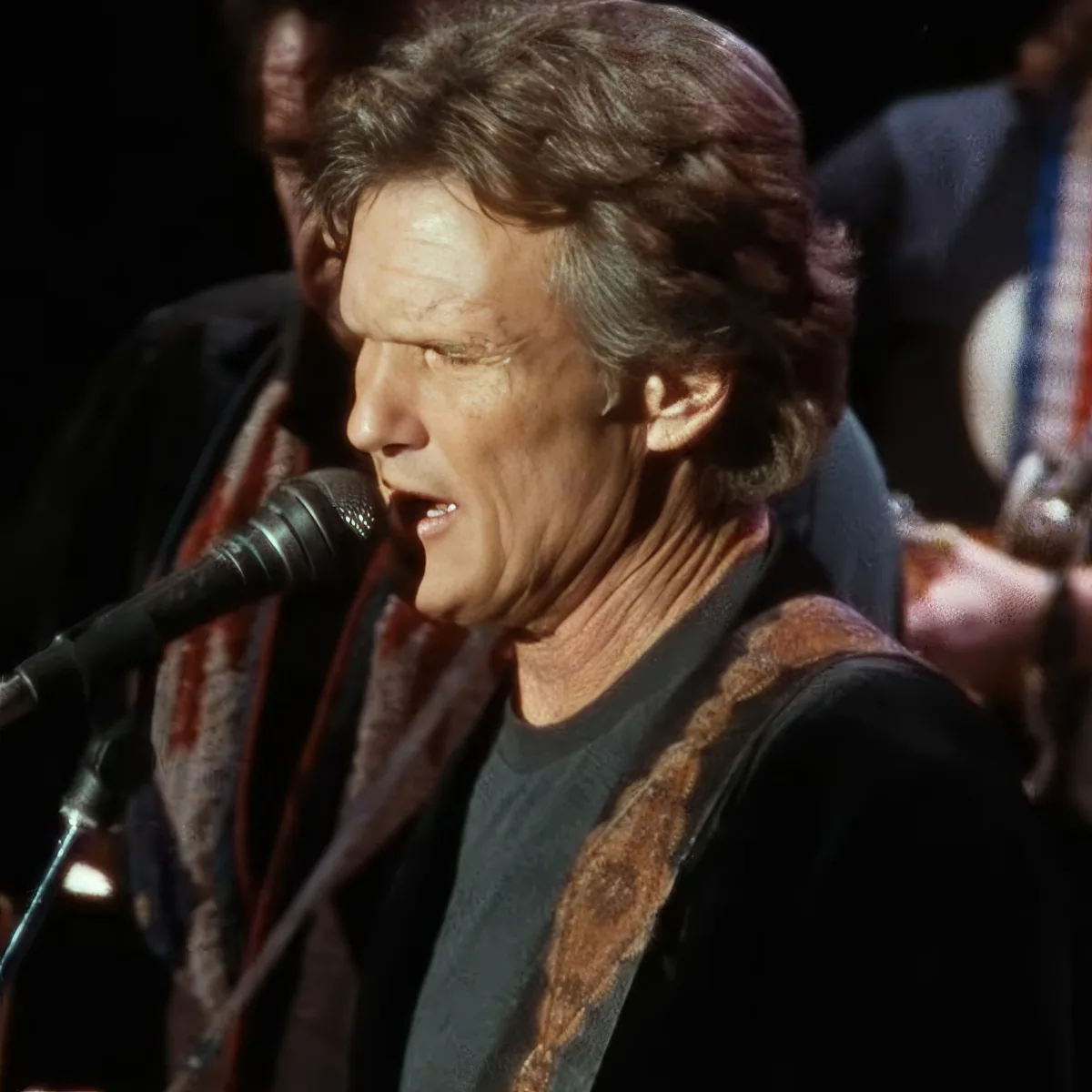 A man with curly hair sings into a microphone, wearing a black shirt and a brown scarf.