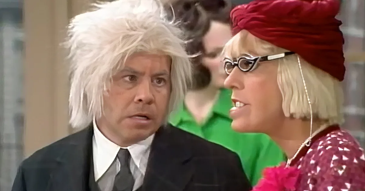 A man with white hair and a suit looks surprised as a woman in a red turban smiles.
