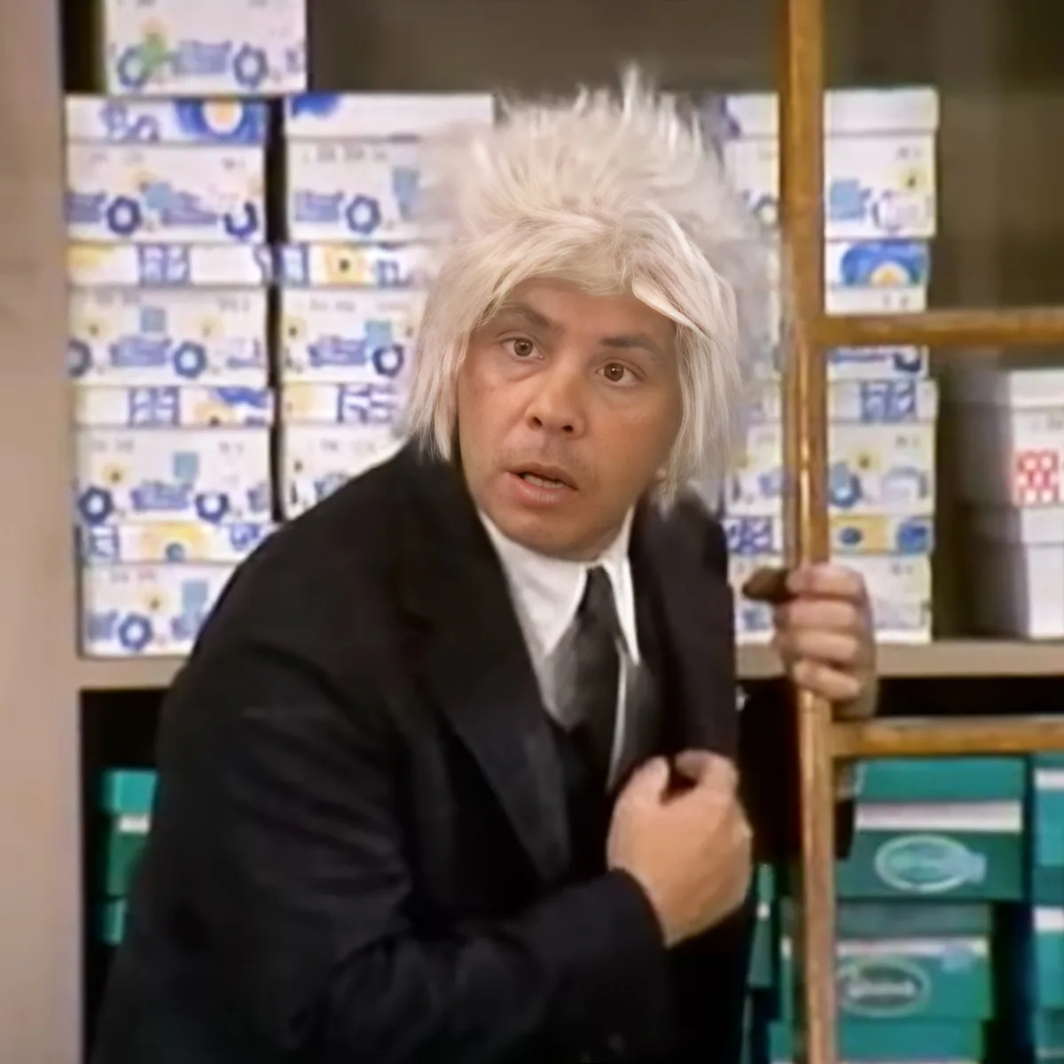 A man with white, spiky hair in a suit holds a cane, standing in front of shelves with boxes.