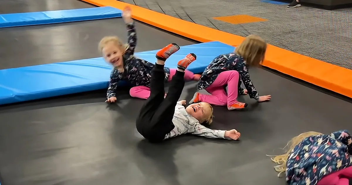 A child in a white shirt and black pants is lying on a trampoline, appearing to be in a playful or possibly distressed...
