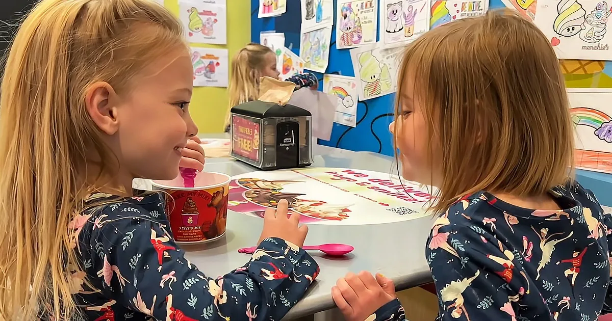 Two young girls in matching pajamas sit at a table, one eating ice cream, surrounded by colorful artwork on the wall.