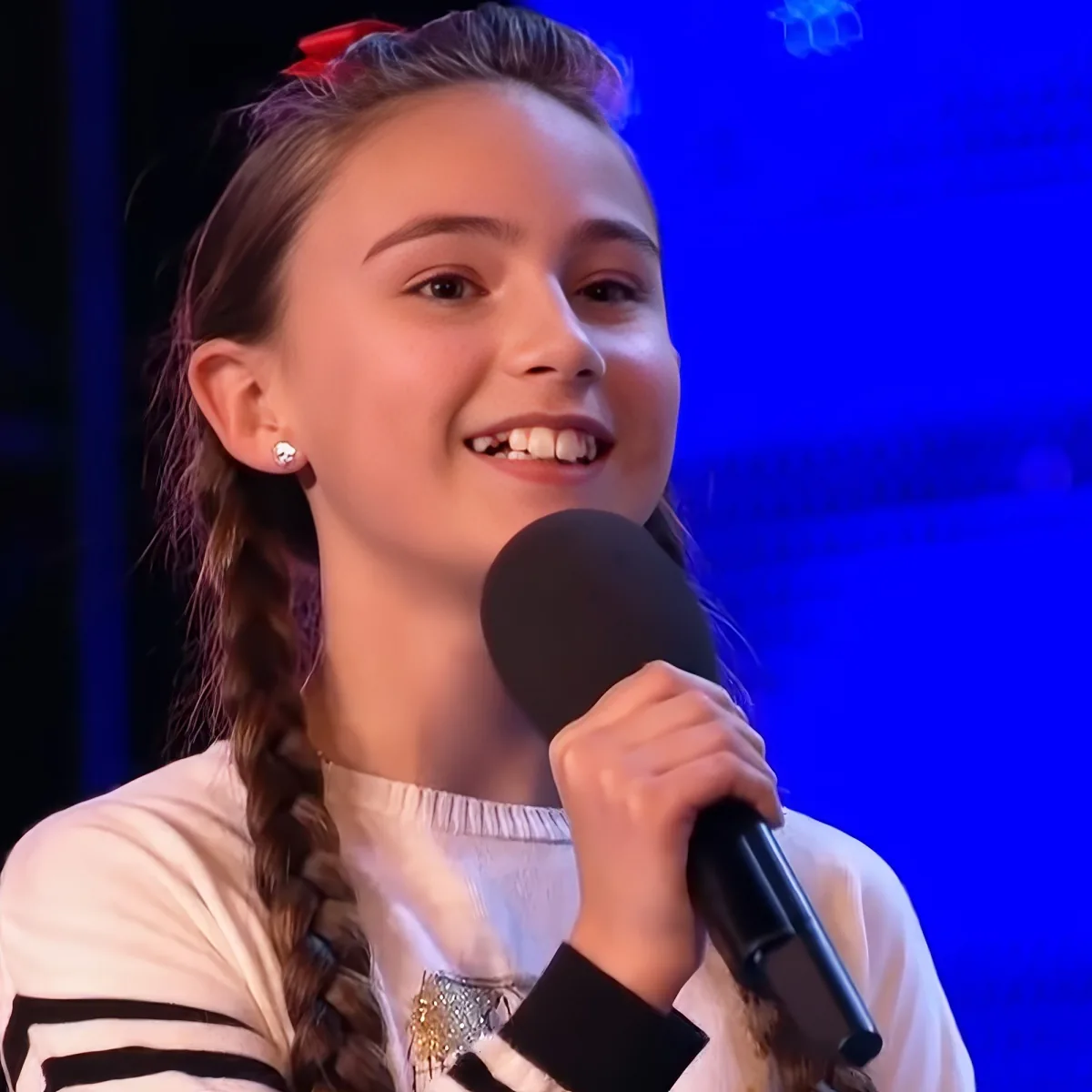 A young girl with braided hair and a red bow smiles while holding a microphone on stage.