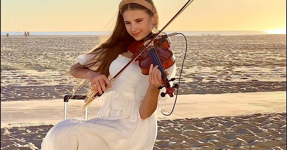 A girl in a white dress plays the violin on a sandy beach at sunset.