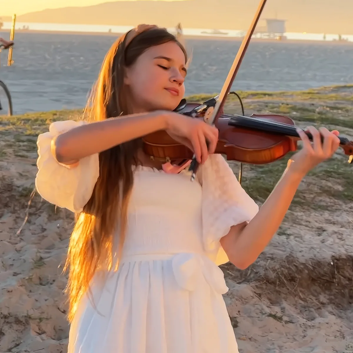 A girl in a white dress plays the violin on a beach at sunset.