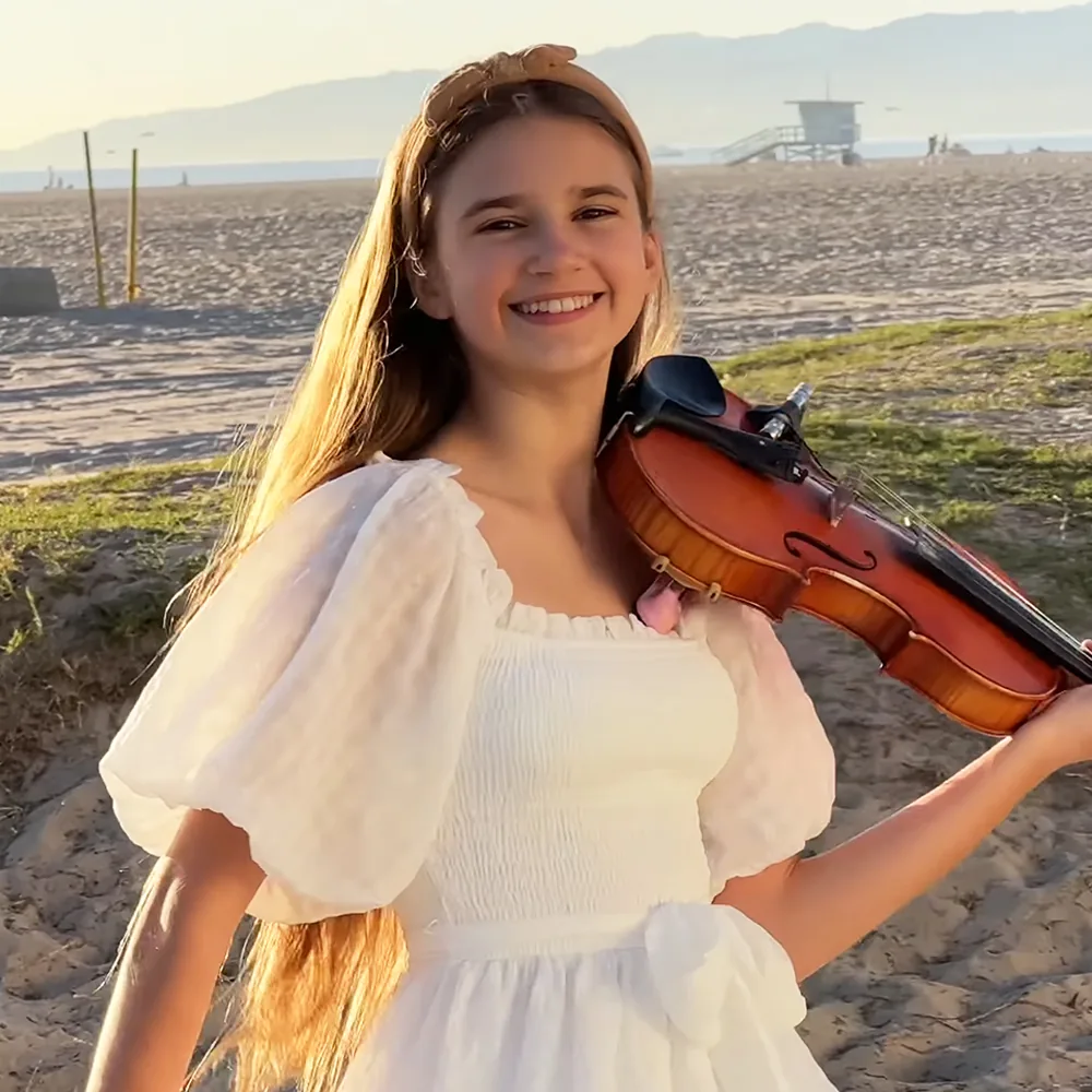 A girl in a white dress smiles while holding a violin on a sandy beach with mountains in the background.