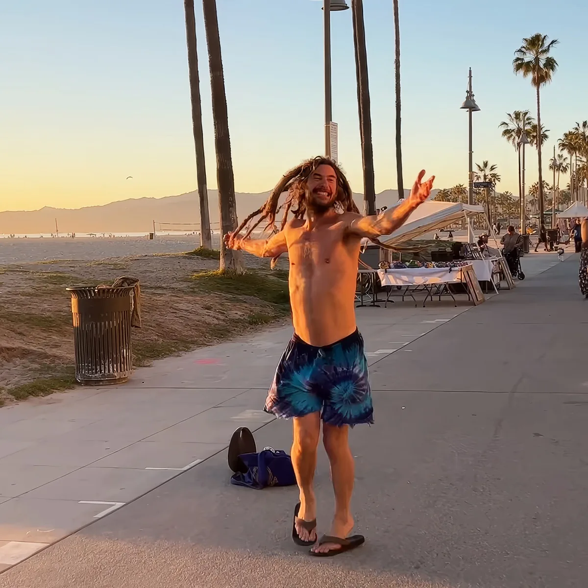A man with dreadlocks and tie-dye shorts dances joyfully on a beachside sidewalk at sunset.