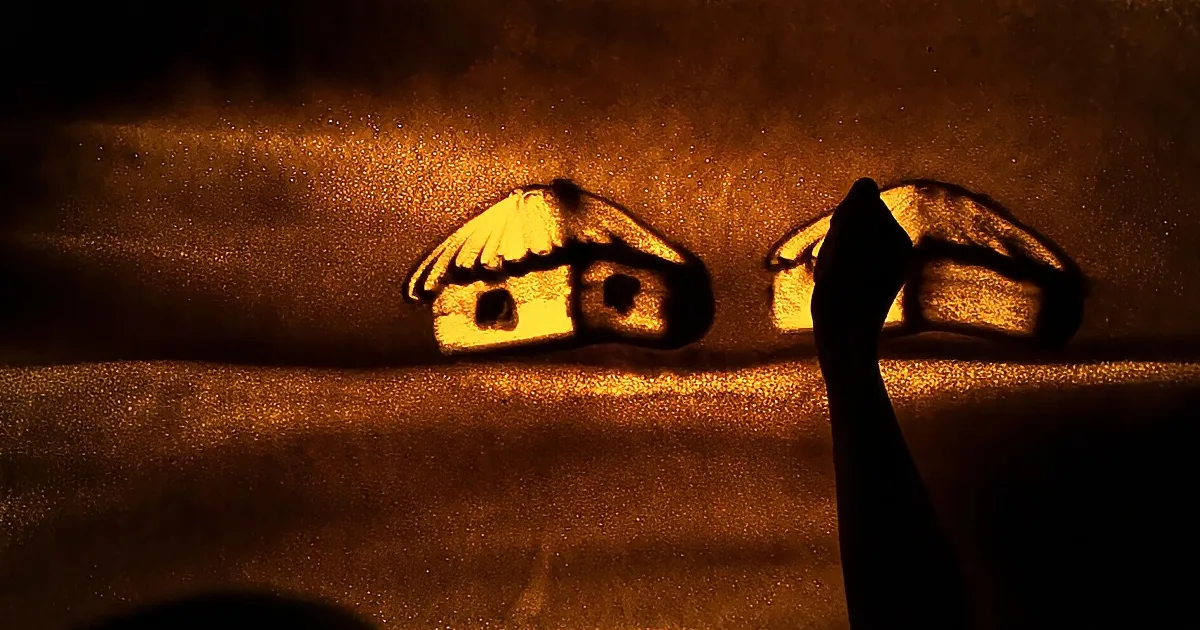 A hand draws a house on a glowing sand surface, creating a warm, glowing scene.