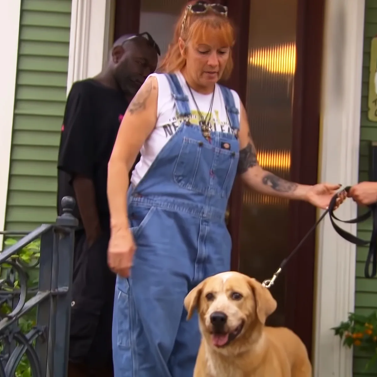 A woman in blue overalls and a white t-shirt is holding a dog leash, with a dog in front of her.