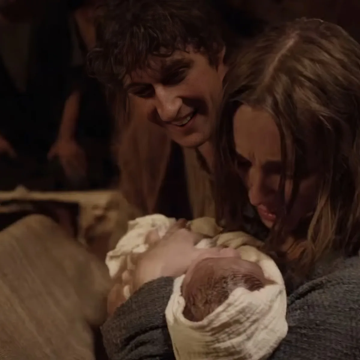 A woman holds a newborn baby wrapped in a blanket, with another person smiling nearby.