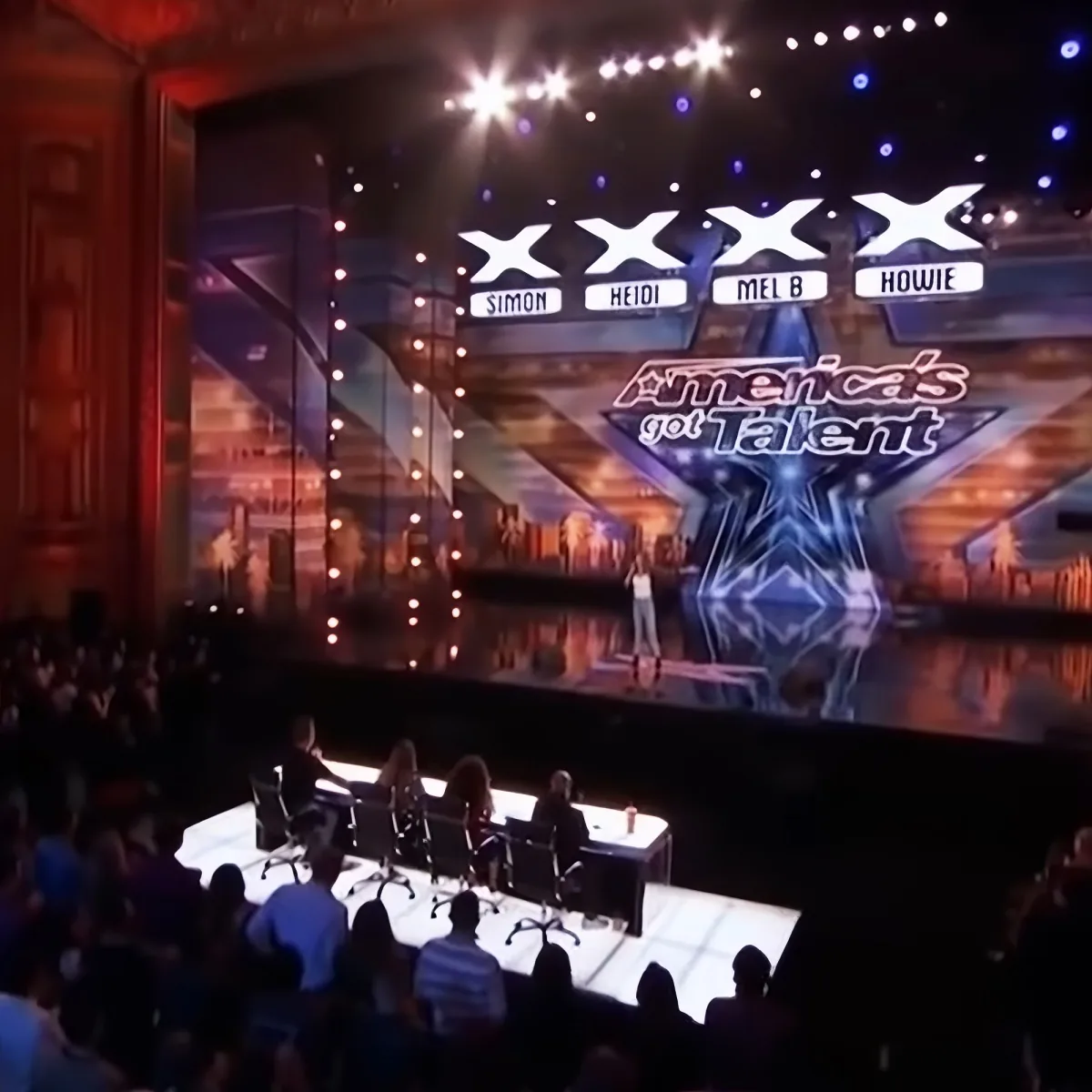 A contestant stands on a brightly lit stage with judges' names displayed above them on "America's Got Talent."