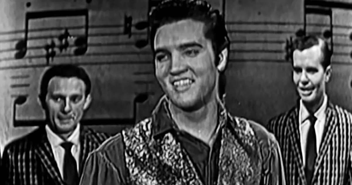 Elvis Presley smiles in a black-and-white scene, wearing a patterned jacket, with two men in striped shirts behind him.