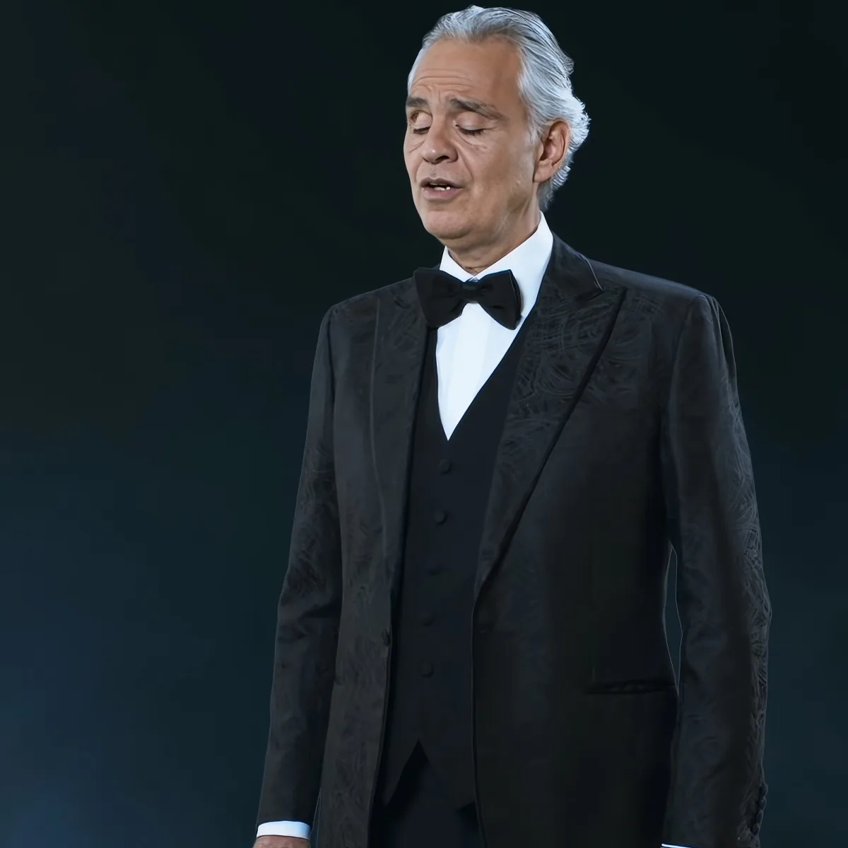 A man in a black tuxedo with a bow tie stands against a dark background, looking downward.