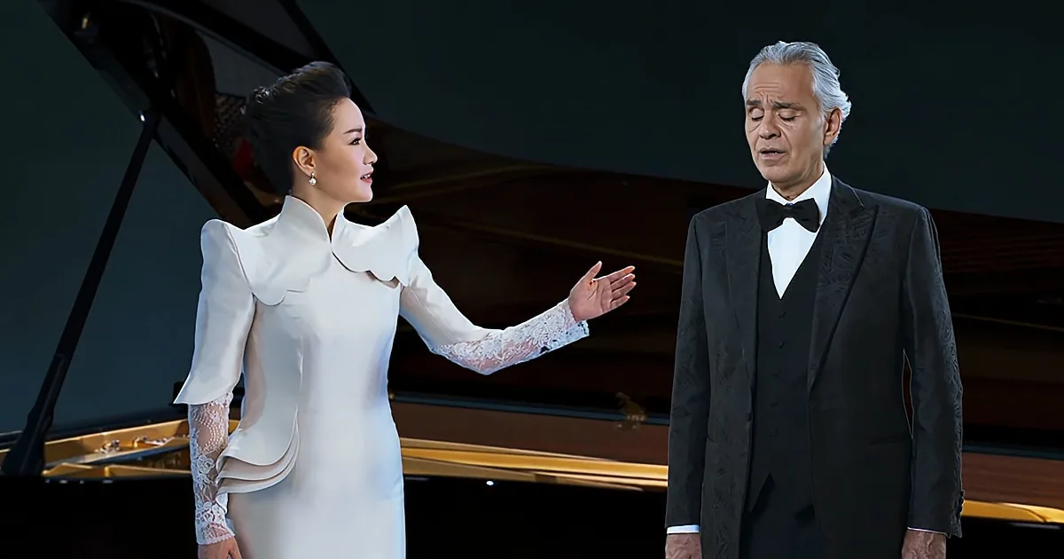 A woman in a white dress gestures towards a man in a black suit standing near a grand piano.