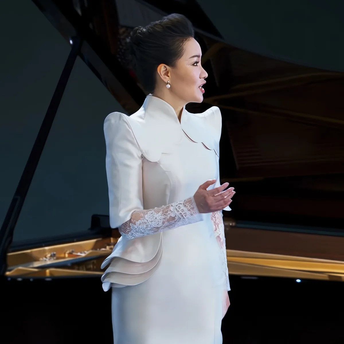 A woman in a white dress with lace sleeves stands in front of a grand piano, singing.