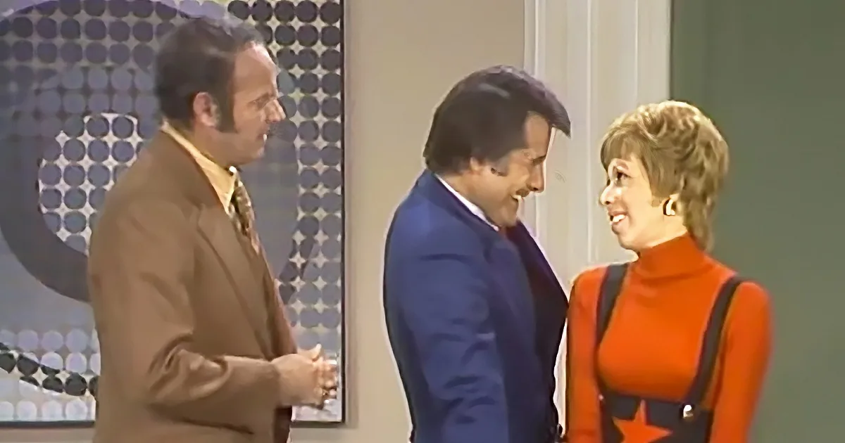 A man in a brown suit and another in a blue suit are engaged in conversation with a woman in an orange outfit.
