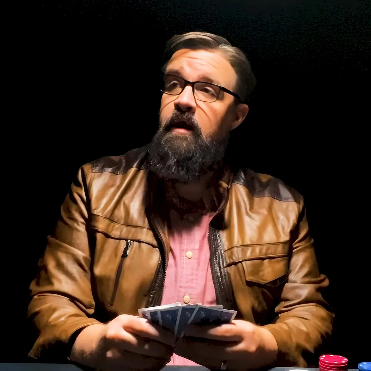 A man with a beard and glasses holds cards, wearing a brown leather jacket and pink shirt, in a dimly lit setting.