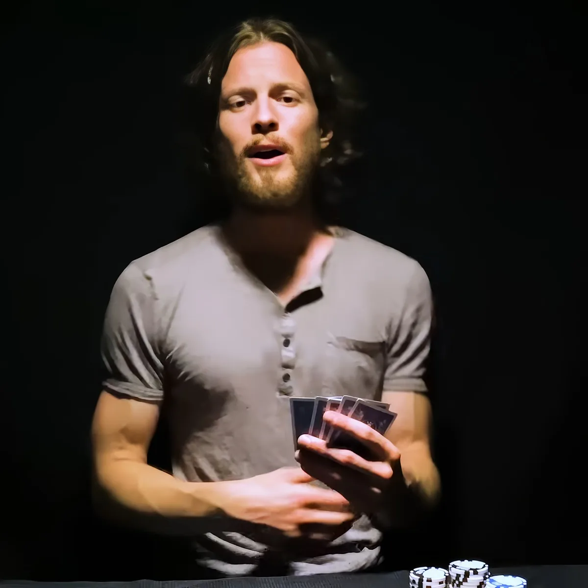 A man in a gray shirt holds a deck of cards, with poker chips visible on a table in the foreground.