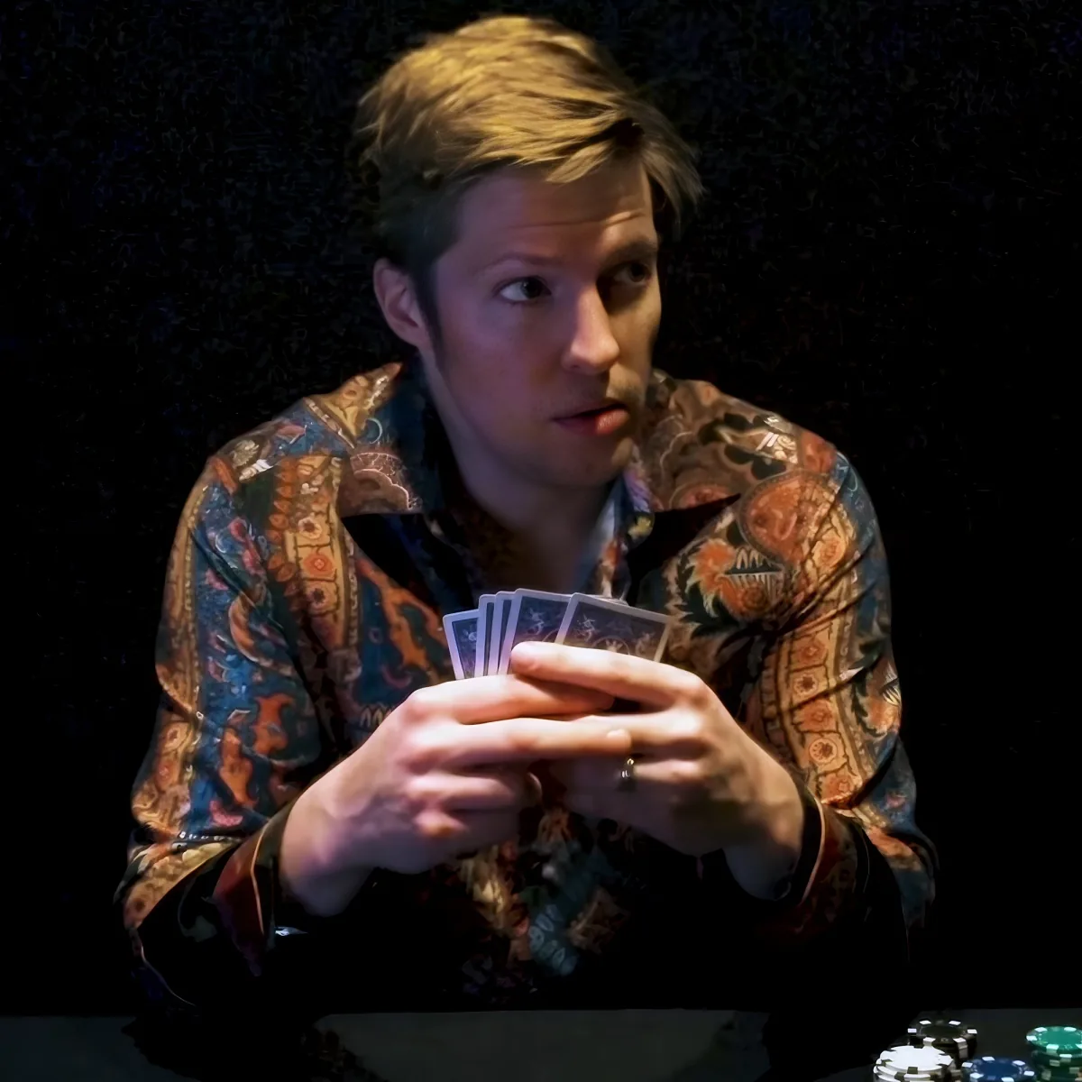 A man in a patterned shirt holds playing cards, looking to the side with a dark background and poker chips visible.