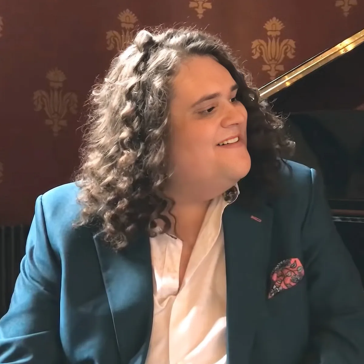 A person with curly hair, wearing a blue blazer and a white shirt, is smiling and looking to the side.