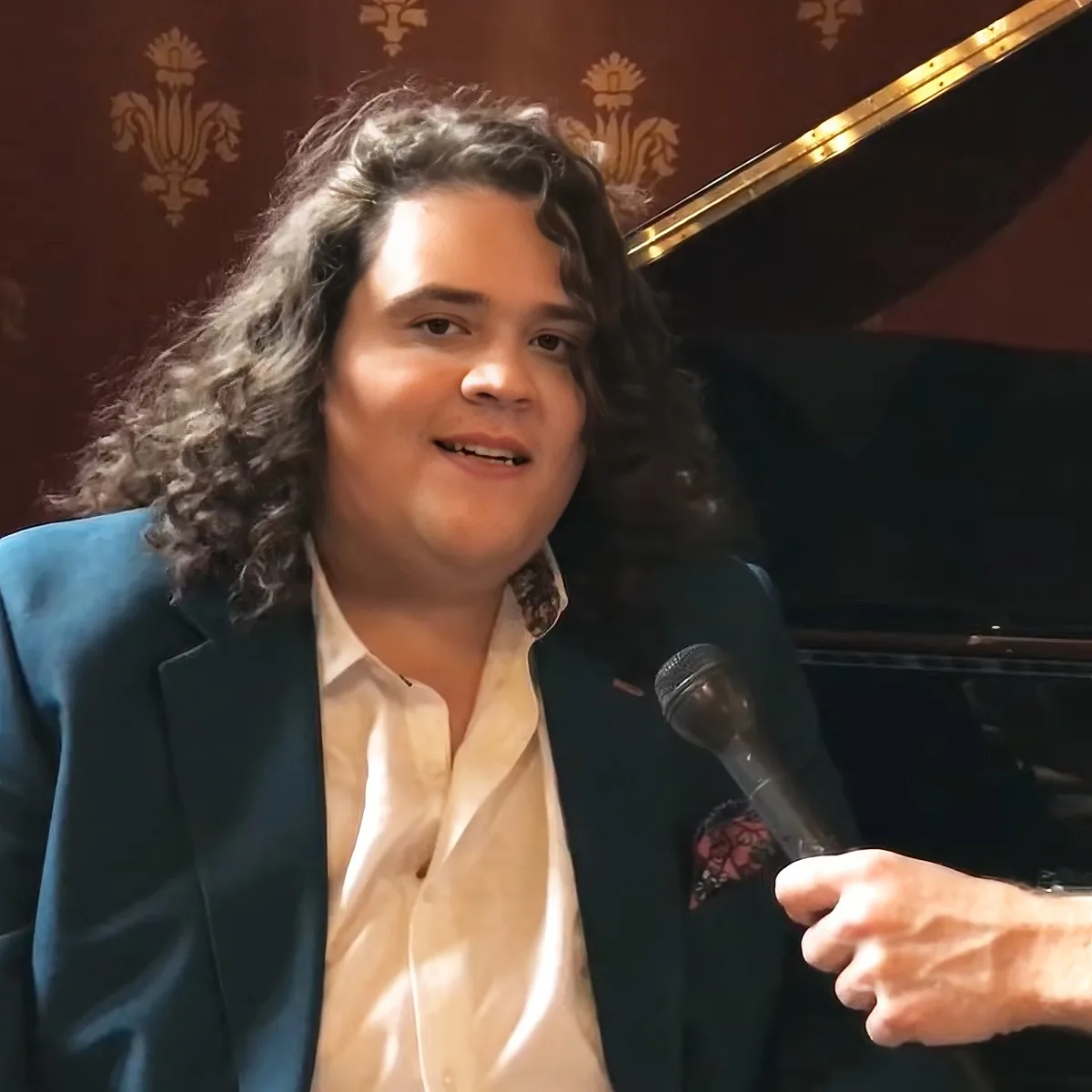 A person with long, curly hair wearing a teal blazer and white shirt is speaking into a microphone.