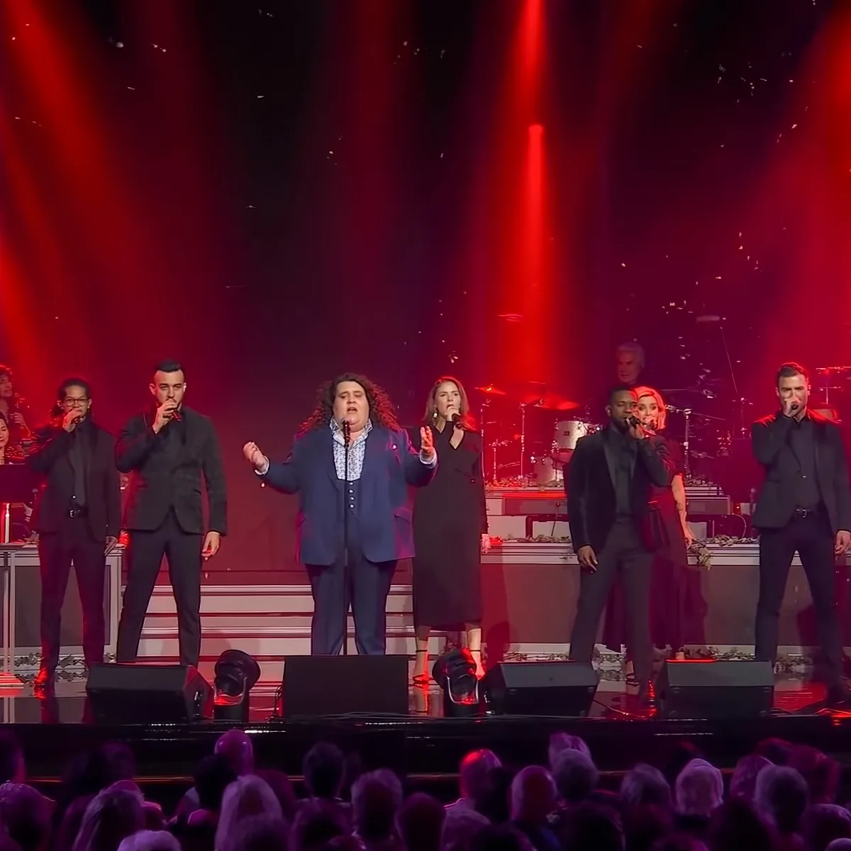 A group of performers, including a man with long hair, sing on stage under red lighting, with a live audience in the...