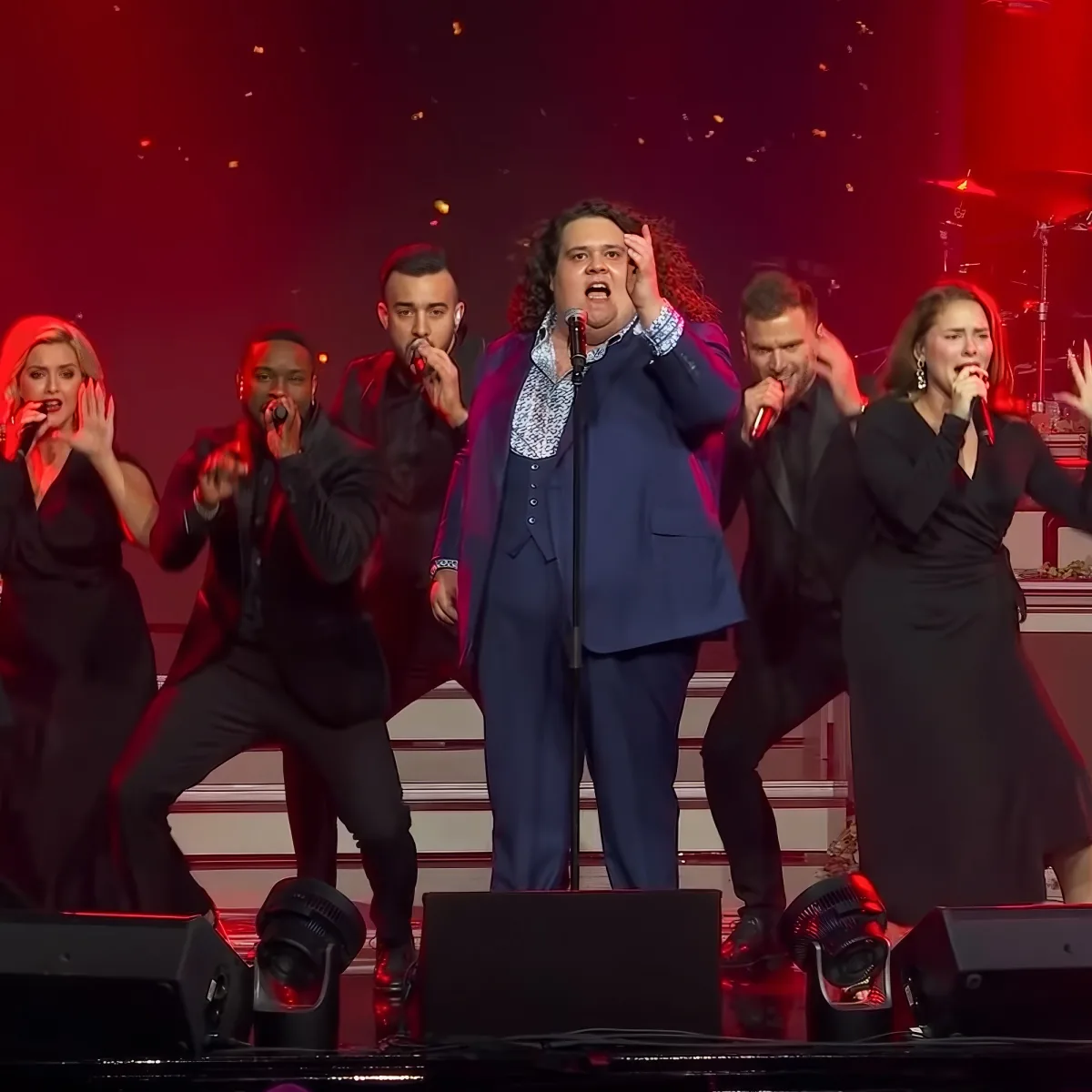 A man in a blue suit sings passionately on stage with a group of performers in black.