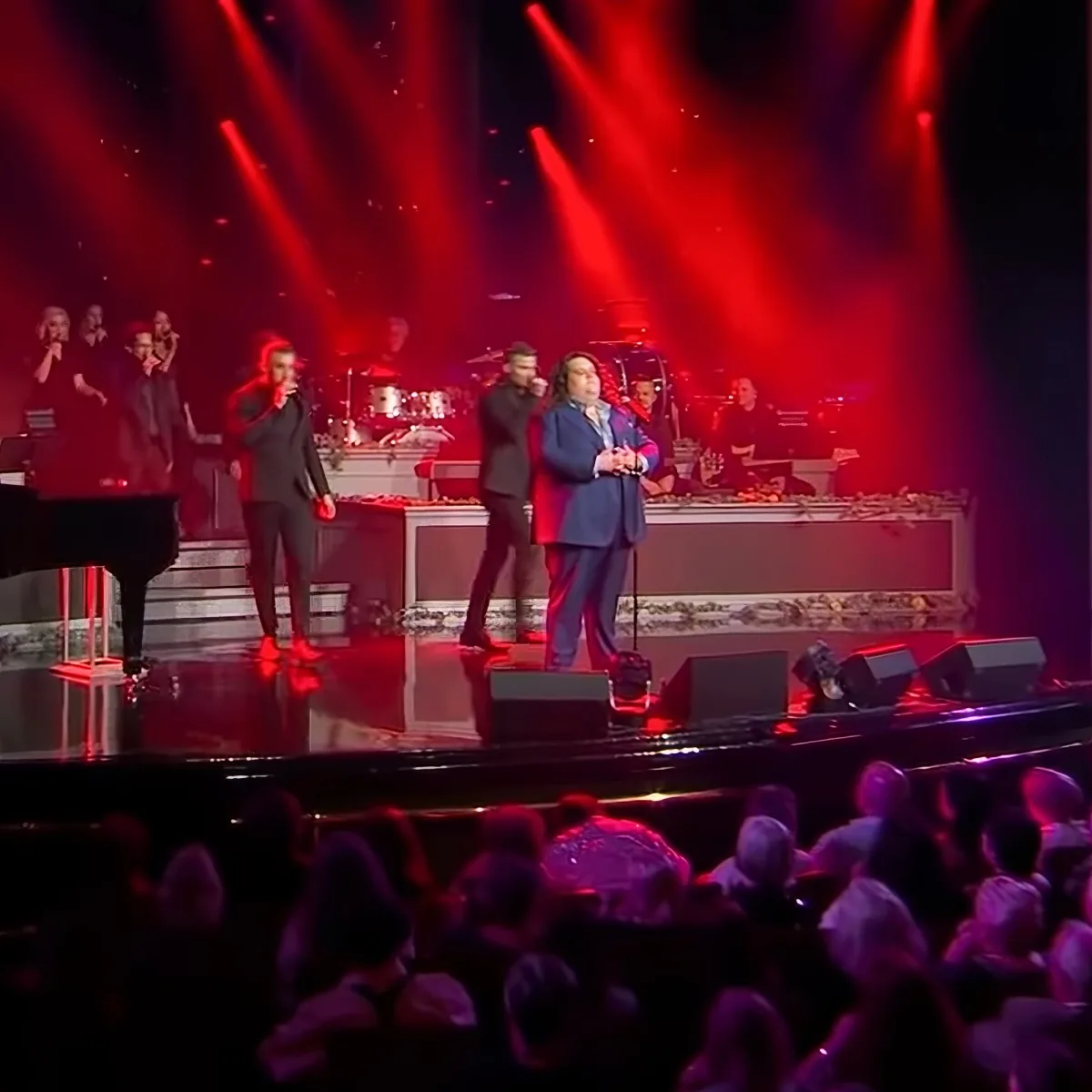 A man in a suit sings on stage with red lights, surrounded by musicians and a piano.